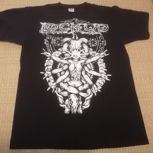 May include: Black t-shirt with a white graphic featuring the band name "LOCK UP" above a detailed illustration of a skeletal figure with multiple arms and a skull. The design is surrounded by a circular pattern and additional text.