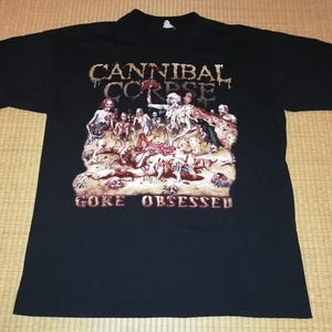 以下が含まれることがあります： 黒いTシャツ。バンド名「Cannibal Corpse」が、使い古されたフォントでグラフィックイラストの上に配置されています。イラストは、人物と「Gore Obsessed」のテキストを含む血なまぐさいシーンを描いています。