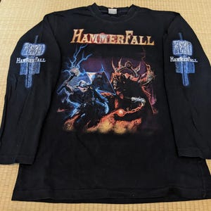 Op de afbeelding: Zwart shirt met lange mouwen met de bandnaam "Hammerfall" in oranje en een afbeelding van twee figuren in gevecht. De mouwen hebben een blauw hamerontwerp met de bandnaam. Het shirt is gemaakt van zacht materiaal.