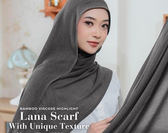 May include: A woman wearing a dark grey Lana scarf with a unique texture. The scarf is made of bamboo viscose and measures 70 cm x 175 cm. The image includes text that says "Lana Scarf With Unique Texture" and "Available in 10+ Colors."
