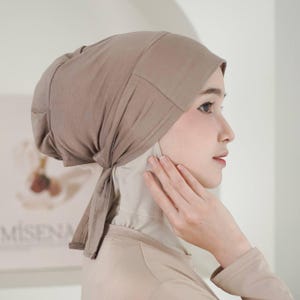 May include: A taupe-colored head covering with a tied back. The head covering is made of a soft, lightweight fabric. The head covering is worn over a light beige under-scarf.