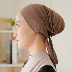 May include: A brown head covering with a gathered back and long ties. The head covering is made of a soft fabric and is worn on the head. The head covering is a solid colour and is designed to cover the hair.