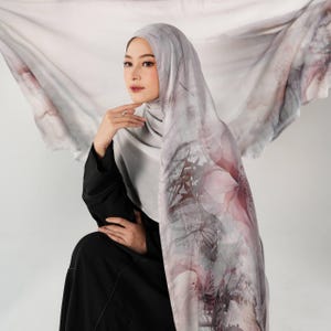 May include: A lightweight headscarf featuring a soft gray base with a watercolor-style floral print in shades of muted pink and dark gray. The fabric drapes around the head and shoulders, showcasing a large, flowing pattern of abstract flower petals.