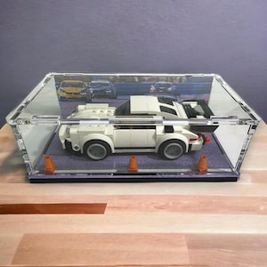 May include: A white Lego sports car model inside a clear acrylic display case. The car has black accents and gray wheels. The case has a black base and orange traffic cones. A racing scene is visible in the background.