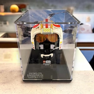May include: A white and orange LEGO Star Wars Luke Skywalker X-Wing Pilot Helmet in a clear acrylic display case with a black base. The base has the LEGO Star Wars logo and the text "STAR WARS".