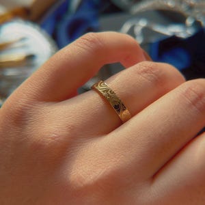 May include: A gold-toned band ring featuring a continuous engraved pattern of swirling lines and organic shapes. The metal surface has a polished finish that reflects light, highlighting the intricate, repeating design etched into the exterior.