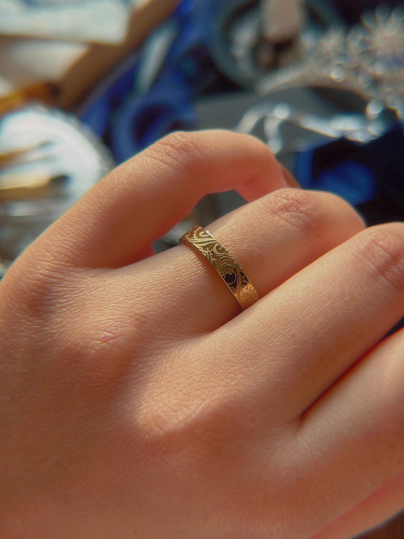 May include: A gold-toned band ring featuring a continuous engraved pattern of swirling lines and organic shapes. The metal surface has a polished finish that reflects light, highlighting the intricate, repeating design etched into the exterior.