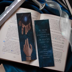 May include: Two fantasy-themed bookmarks with dark blue backgrounds and illustrations of hands and text. A silver sword and open book are also visible, creating a mystical aesthetic.
