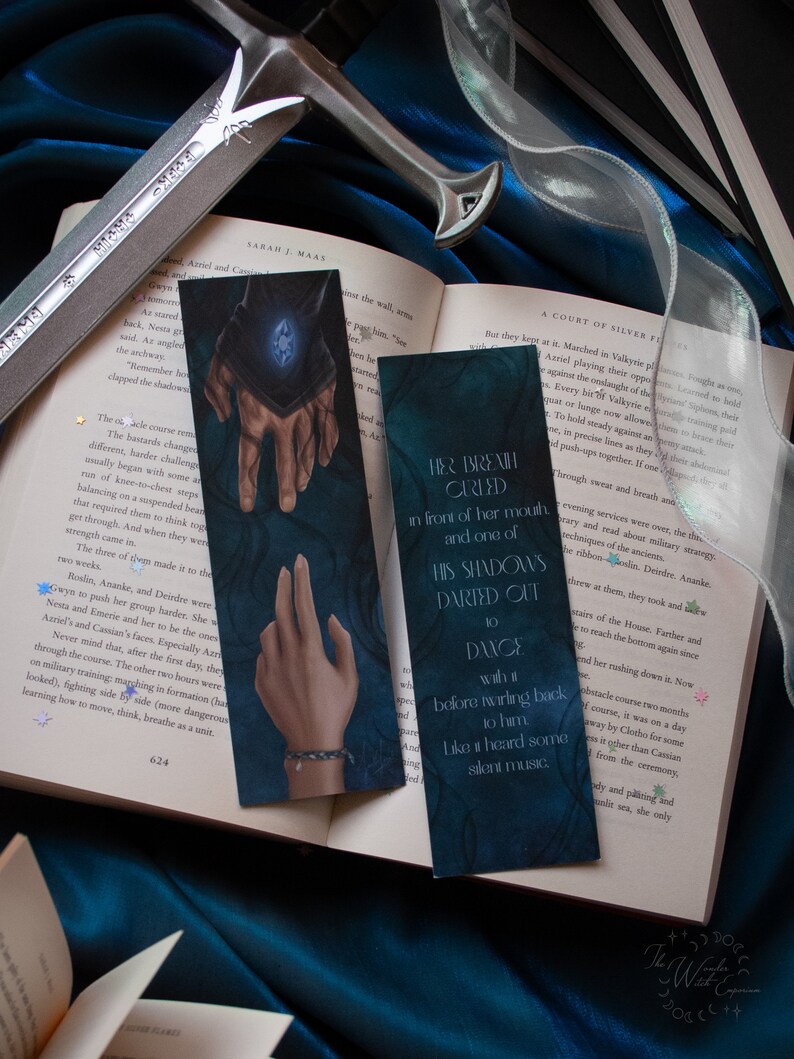 May include: Two fantasy-themed bookmarks with dark blue backgrounds and illustrations of hands and text. A silver sword and open book are also visible, creating a mystical aesthetic.