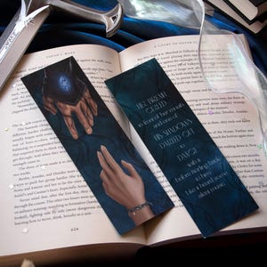 May include: Two fantasy-themed bookmarks with illustrations of hands and text, resting on an open book. A silver sword and ribbon are also visible. The bookmarks have a dark blue background with white text.