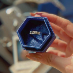 May include: A silver-toned metal band ring featuring an intricate, engraved swirling pattern across its surface. The ring is displayed inside a deep blue, hexagonal-shaped velvet jewelry box with a matching blue interior lining.