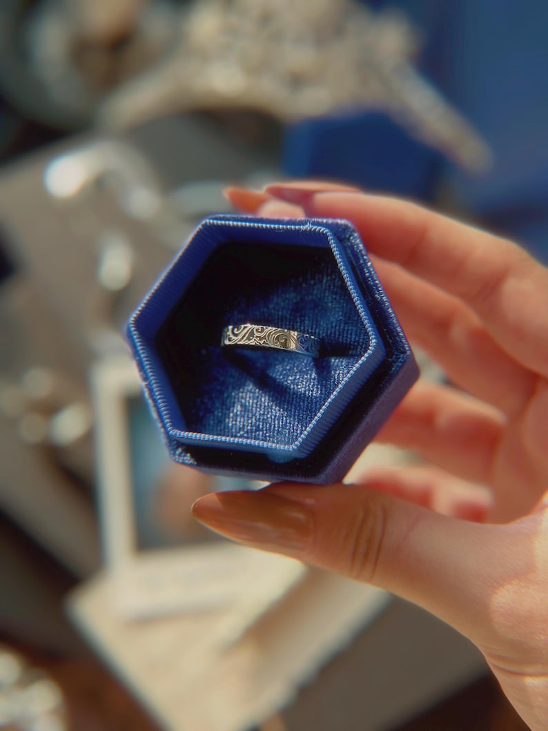 May include: A silver-toned metal band ring featuring an intricate, engraved swirling pattern across its surface. The ring is displayed inside a deep blue, hexagonal-shaped velvet jewelry box with a matching blue interior lining.