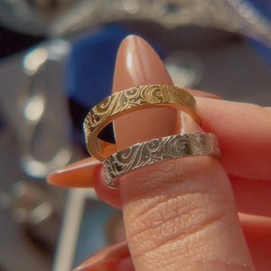 May include: Two metal rings, one gold-toned and one silver-toned, each featuring a continuous embossed swirling floral pattern across the outer surface. The bands have a flat profile and a polished finish, showcasing intricate engraved details.
