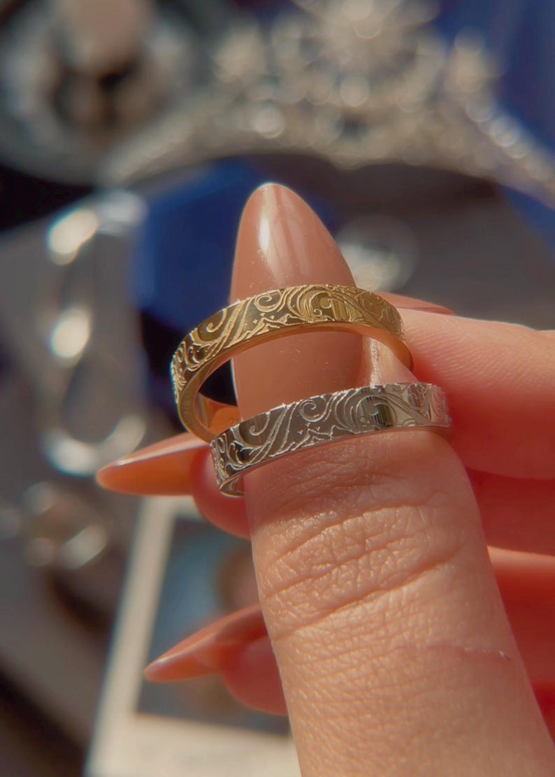 May include: Two metal rings, one gold-toned and one silver-toned, each featuring a continuous embossed swirling floral pattern across the outer surface. The bands have a flat profile and a polished finish, showcasing intricate engraved details.