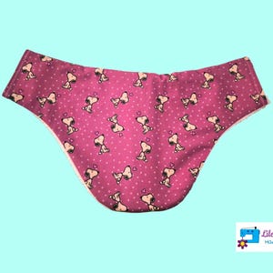 Può includere: Intimo rosa con bordino bianco e un motivo ripetuto di cani dei cartoni animati con cuori. Il testo "Lilo Telitas Hand Made" è visibile nell'angolo in basso a destra.