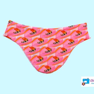 May include: Pink fabric underwear with a repeating pattern of cartoon girls with orange hair and pink dresses. The fabric has a glittery texture.