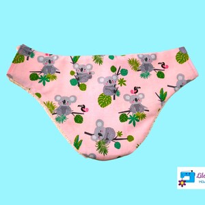 Peut inclure: Bavoir bébé rose avec un imprimé koala et toucan. Le bavoir a une bordure blanche et dit "Lilo Telitas Hand Made" dans le coin inférieur droit.