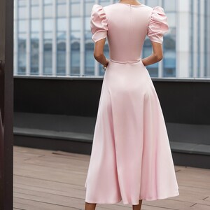 A light pink midi dress with short, puffed sleeves and a flared skirt. The dress features a fitted bodice and a subtle waistline. The model is wearing white heels. The background includes a modern building.