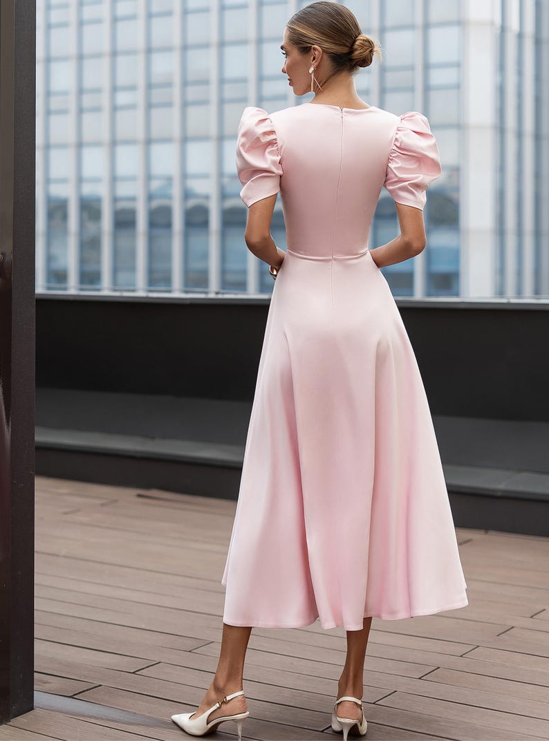 A light pink midi dress with short, puffed sleeves and a flared skirt. The dress features a fitted bodice and a subtle waistline. The model is wearing white heels. The background includes a modern building.