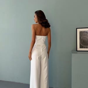 May include: A strapless white jumpsuit with wide-leg pants. The outfit features a fitted bodice and a zipper closure down the back. The model is standing in front of a light blue wall.