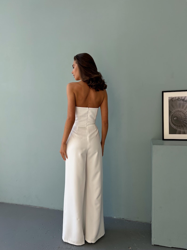May include: A strapless white jumpsuit with wide-leg pants. The outfit features a fitted bodice and a zipper closure down the back. The model is standing in front of a light blue wall.