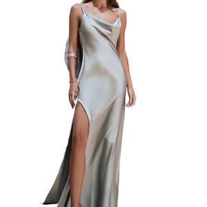 May include: A flowing, pale green satin gown with a cowl neckline and a high slit. The dress features thin straps and a draped back detail. The model is wearing gold earrings and sandals.