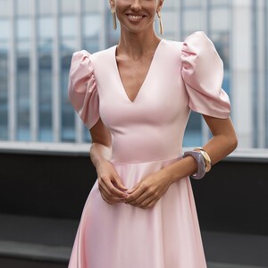 A light pink dress with a V-neckline and short, puffed sleeves. The dress has a flowing skirt and is accessorized with gold earrings and bracelets. The background features a modern building.