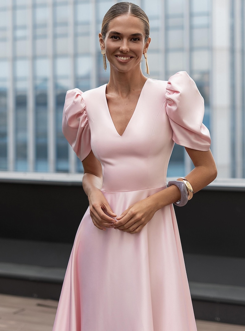 A light pink dress with a V-neckline and short, puffed sleeves. The dress has a flowing skirt and is accessorized with gold earrings and bracelets. The background features a modern building.