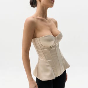 May include: A strapless, cream-colored corset with a structured bodice and a peplum hem. The corset features vertical paneling and a smooth, satin-like finish. The model is wearing black leggings.