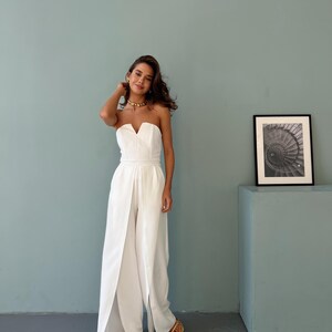 May include: A strapless white jumpsuit with a V-neckline and wide-leg design. The jumpsuit features a split front, creating a flowing silhouette. The model is wearing gold sandals and a gold necklace. A framed black and white photo is on the right.