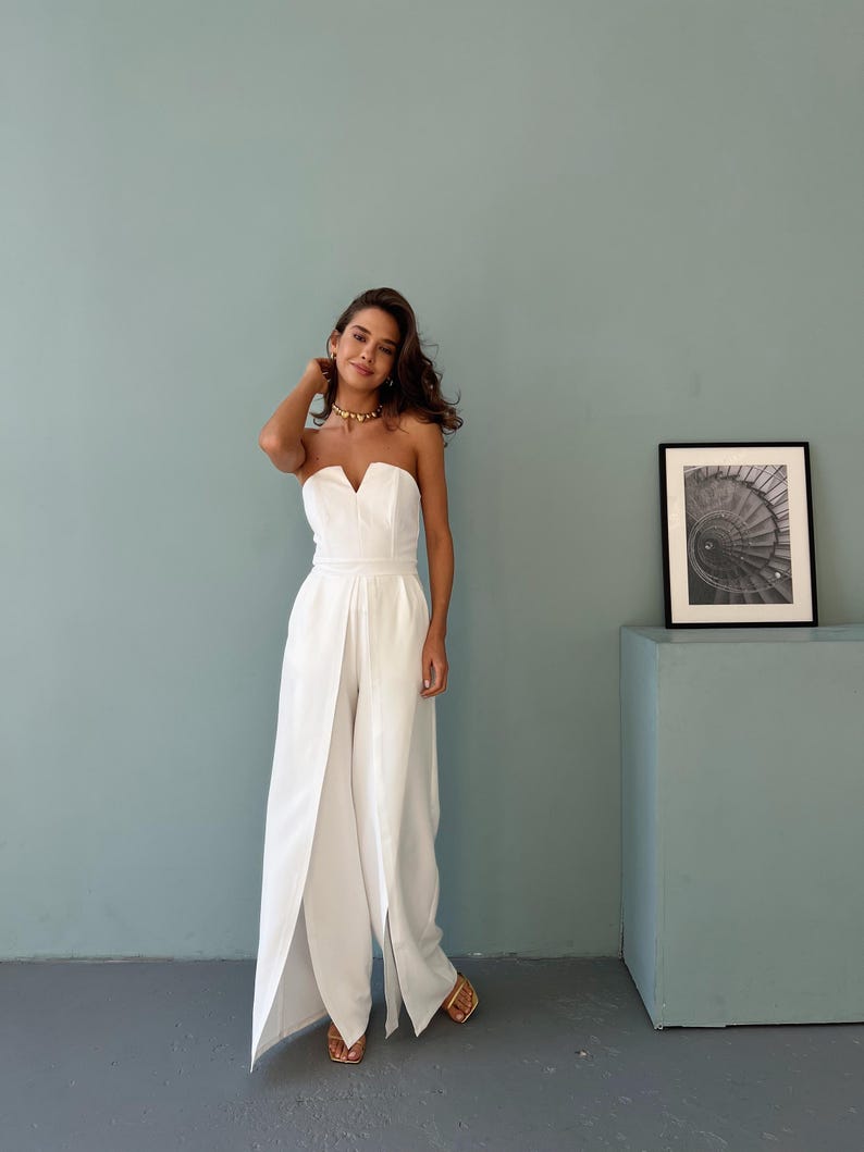 May include: A strapless white jumpsuit with a V-neckline and wide-leg design. The jumpsuit features a split front, creating a flowing silhouette. The model is wearing gold sandals and a gold necklace. A framed black and white photo is on the right.