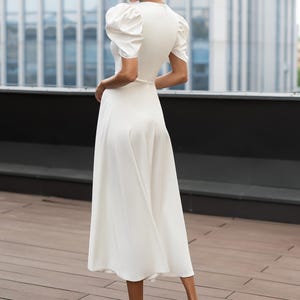 Back full length view of a white midi dress with short, puffed sleeves and a v-neckline. The dress has a fitted bodice and a flowing skirt. The model is wearing white heels. The dress is suitable for formal events.