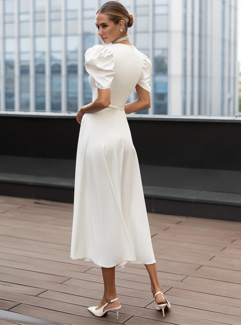 Back full length view of a white midi dress with short, puffed sleeves and a v-neckline. The dress has a fitted bodice and a flowing skirt. The model is wearing white heels. The dress is suitable for formal events.