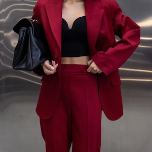 May include: A burgundy pantsuit with a matching blazer and trousers. The blazer has a classic cut with a two-button closure. A black top and a black handbag complete the outfit. The background is a metallic wall.