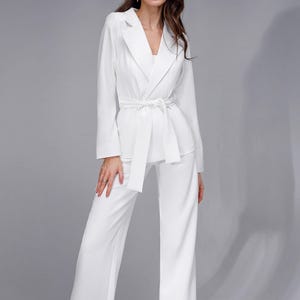 May include: A solid white two-piece suit consisting of a long-sleeved blazer with a notched lapel and a matching fabric tie belt at the waist, paired with straight-leg trousers. The blazer features a clean, structured design with long sleeves.