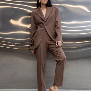 Pu&ograve; includere: Un completo marrone con blazer e pantaloni abbinati. Il blazer ha revers a lancia e chiusura a un bottone. I pantaloni sono a gamba dritta. La persona indossa scarpe slip-on in camoscio marrone. Lo sfondo &egrave; una parete metallica.