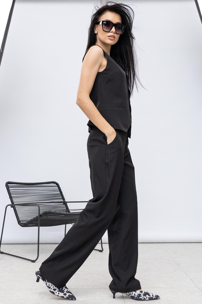 May include: A black sleeveless vest and wide-leg trousers suit. The outfit is paired with black and white patterned heels and large sunglasses. A black wire chair is in the background.