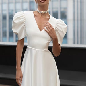 A white satin dress with a V-neckline and short, puffed sleeves. The dress features a fitted bodice and a flowing skirt. The model is wearing a pearl choker necklace and pearl earrings. The background is a blurred cityscape.