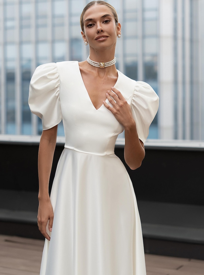 A white satin dress with a V-neckline and short, puffed sleeves. The dress features a fitted bodice and a flowing skirt. The model is wearing a pearl choker necklace and pearl earrings. The background is a blurred cityscape.