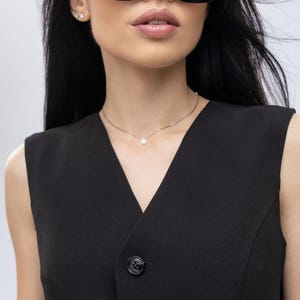 May include: A black sleeveless waistcoat with two buttons is shown. The waistcoat is paired with large, square-framed sunglasses with gradient lenses. A delicate silver necklace with a single pendant is also visible.