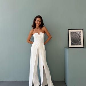 May include: A white strapless jumpsuit with a sweetheart neckline and wide-leg design. The jumpsuit features a fitted bodice and a high waist, with a split front. The model is wearing gold-colored jewelry and sandals. The background is a light blue wall.