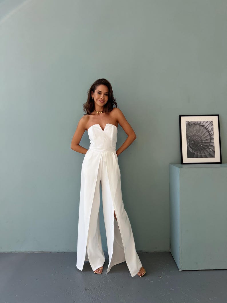 May include: A white strapless jumpsuit with a sweetheart neckline and wide-leg design. The jumpsuit features a fitted bodice and a high waist, with a split front. The model is wearing gold-colored jewelry and sandals. The background is a light blue wall.