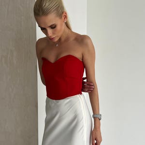 May include: A strapless red bustier top paired with a white satin skirt. The top has a sweetheart neckline. The skirt is a midi length with a side zipper. The outfit is a chic, elegant ensemble.