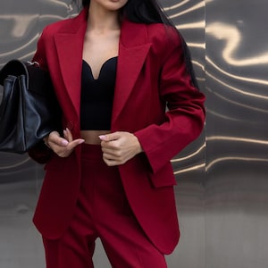 May include: A burgundy pantsuit with a matching blazer and trousers. The blazer has a classic lapel and a single-button closure. A black crop top is worn underneath. A black handbag is held in the left hand.