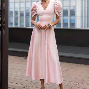 A light pink midi dress with a V-neckline and short, puffed sleeves. The dress has a fitted bodice and a flowing skirt. The model is wearing white heels. The background features a modern building.