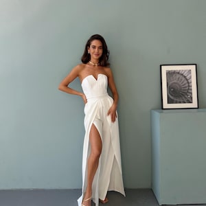 May include: A white strapless jumpsuit with a sweetheart neckline and high slits on the legs. The model is wearing gold jewelry and sandals. The background is a light blue wall and a gray floor.