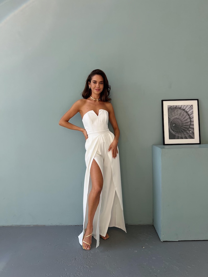 May include: A white strapless jumpsuit with a sweetheart neckline and high slits on the legs. The model is wearing gold jewelry and sandals. The background is a light blue wall and a gray floor.