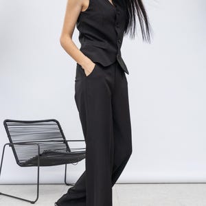 May include: Black waistcoat and wide-leg trousers suit. The waistcoat has a V-neck and button closure. The trousers are high-waisted. The shoes have a black and white pattern. A black chair is in the background.