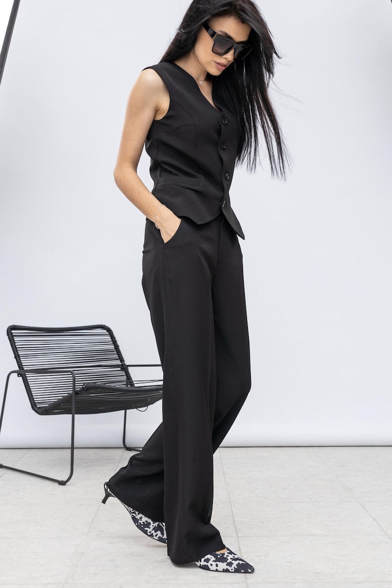 May include: Black waistcoat and wide-leg trousers suit. The waistcoat has a V-neck and button closure. The trousers are high-waisted. The shoes have a black and white pattern. A black chair is in the background.