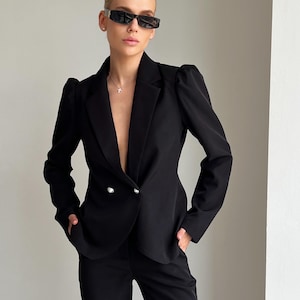 Könnte beinhalten: Ein schwarzer Blazer mit tailliertem Schnitt, doppelreihigem Verschluss mit silbernen Knöpfen und einem fallenden Revers. Der Blazer hat strukturierte Schultern und wird mit einer passenden schwarzen Hose kombiniert. Die Person trägt eine schwarze Sonnenbrille.